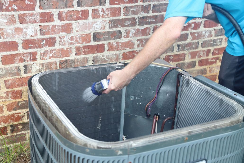 Person cleaning an air conditioning unit with a hose against a brick wall.