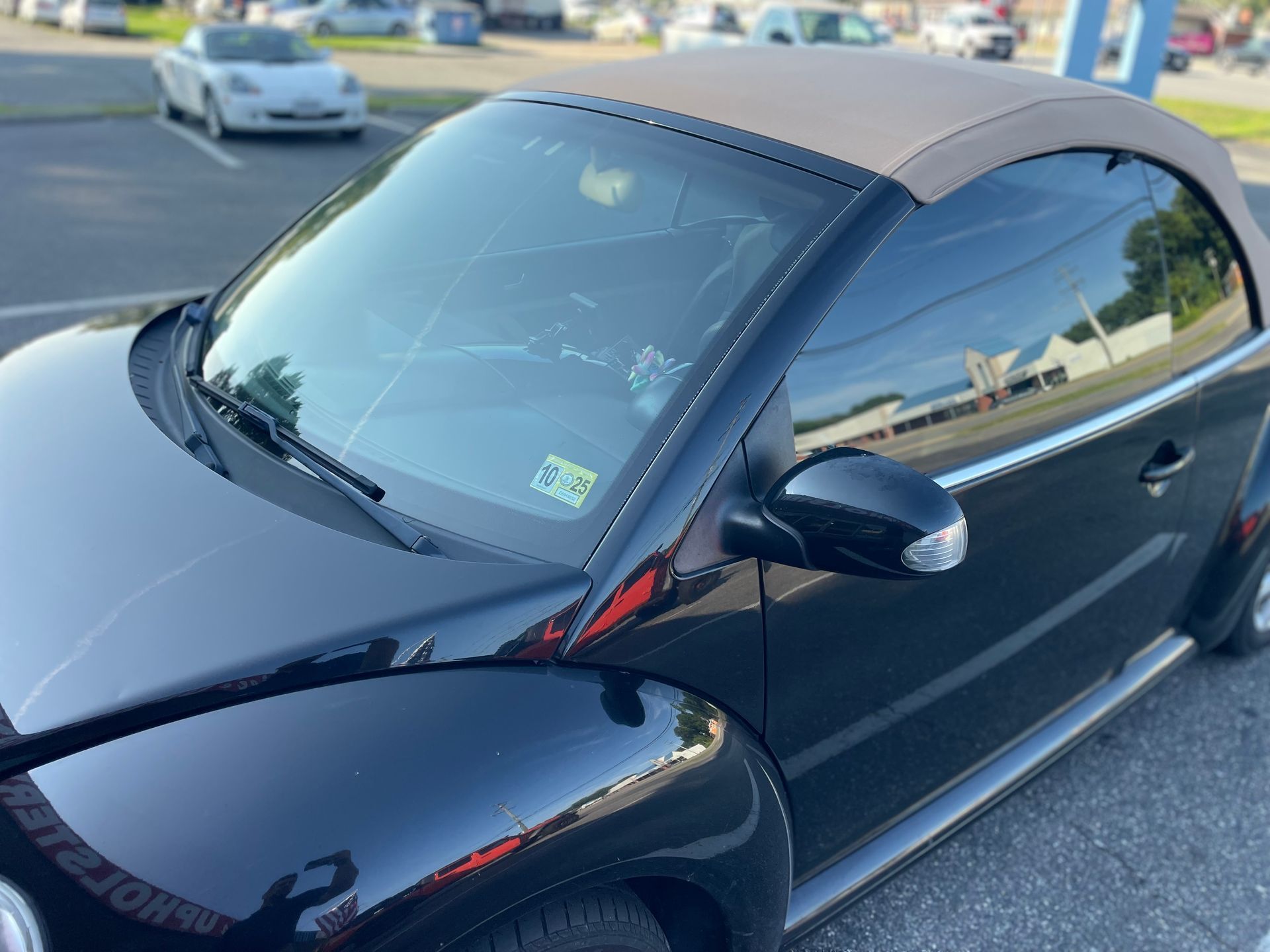Black Volkswagen Beetle convertible with a tan top, parked outside on a sunny day. The windshield has cracks.