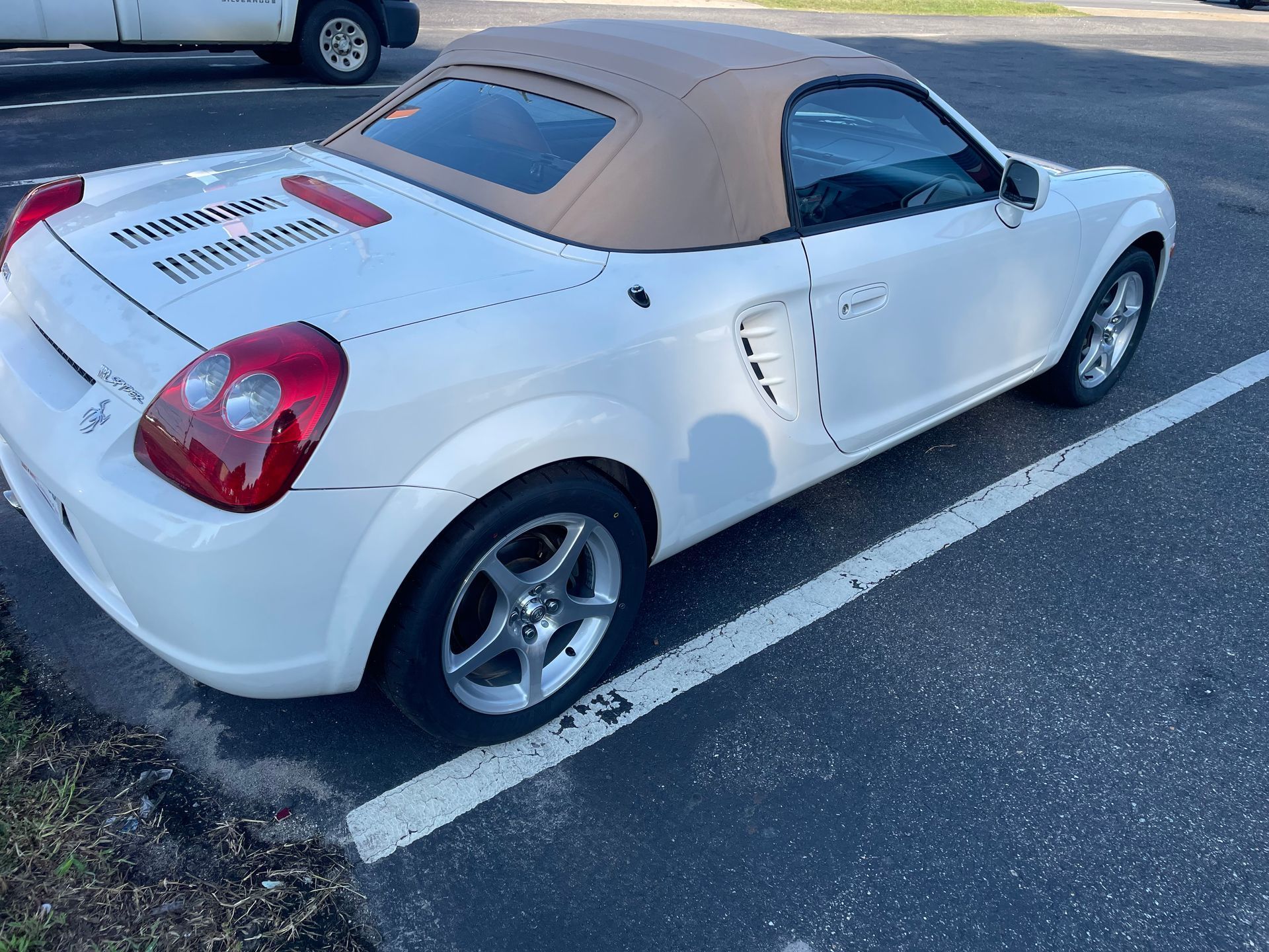 White Toyota MR2 Spyder convertible with a tan soft top parked on asphalt next to a curb.