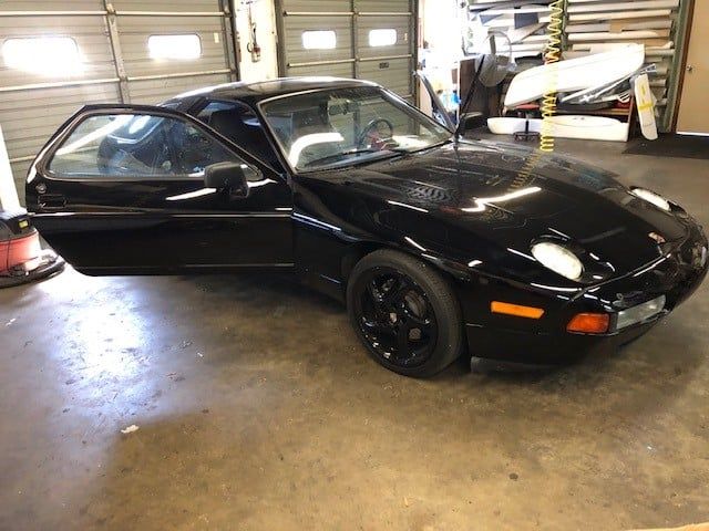 A black porsche 928 is parked in a garage with its doors open.