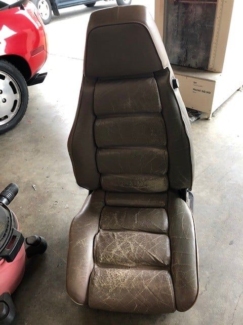 A brown leather car seat is sitting in a garage next to a red car
