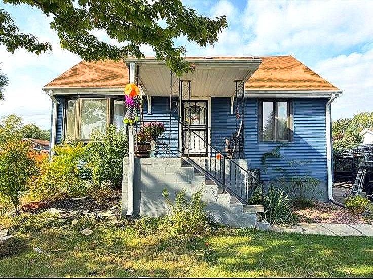 Blue house with a brown shingled roof, a front porch, concrete steps, and a grassy lawn under a sunny sky.