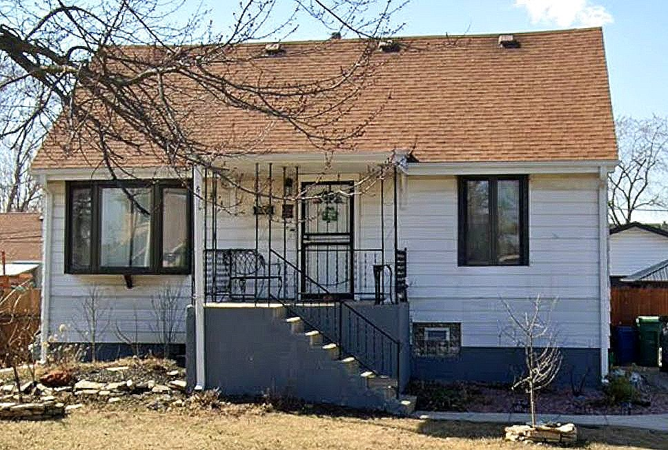A one-story white house with a brown shingled roof, a front porch with black metal railings, and concrete stairs.