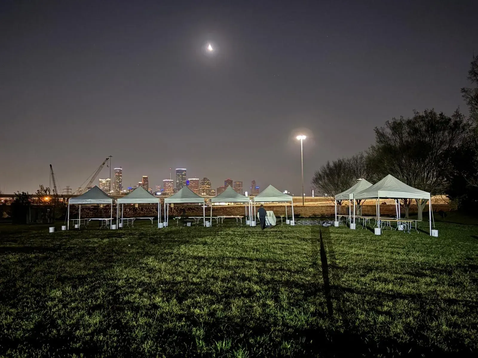 Tents set up on a grassy field at night, cityscape in the background, a crescent moon visible.