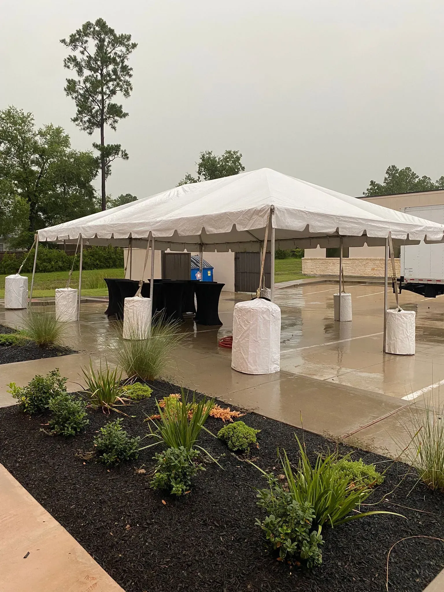 White tent set up outdoors in the rain over tables and equipment, with plants in foreground and cloudy sky.