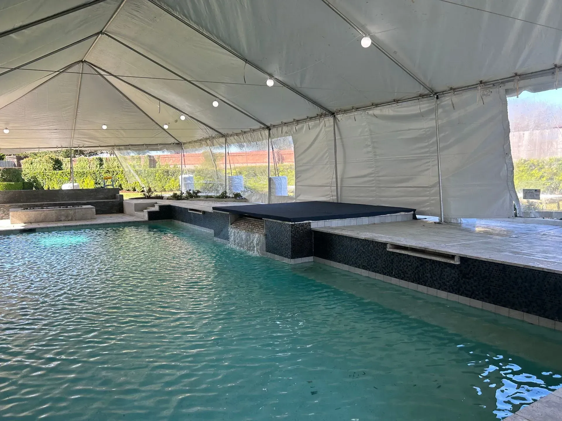 Pool under a white tent. Turquoise water, dark tile trim, and string lights. Sunny outdoors.