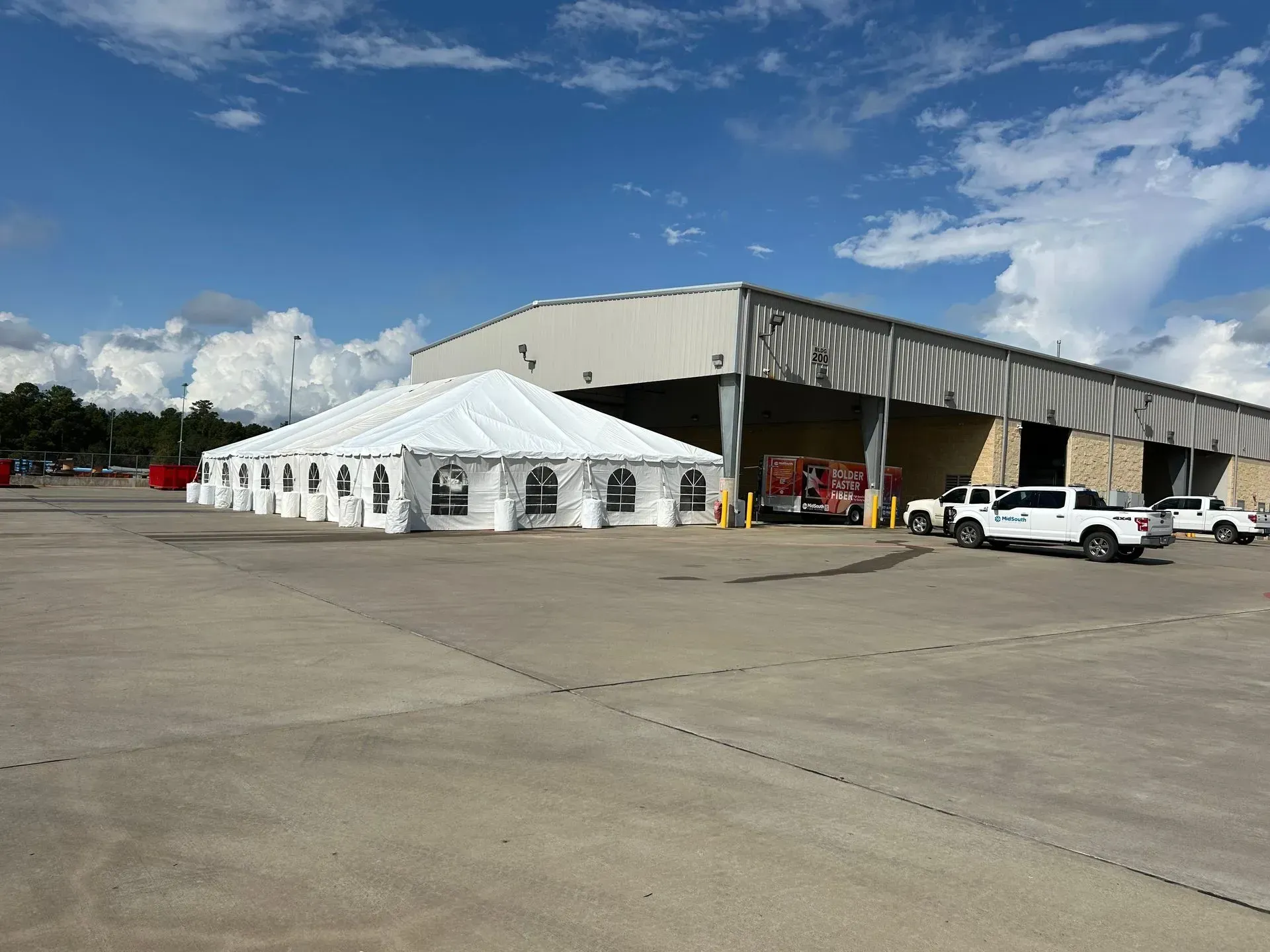 White tent set up next to a large warehouse, with vehicles and a fire truck parked nearby under a partly cloudy sky.