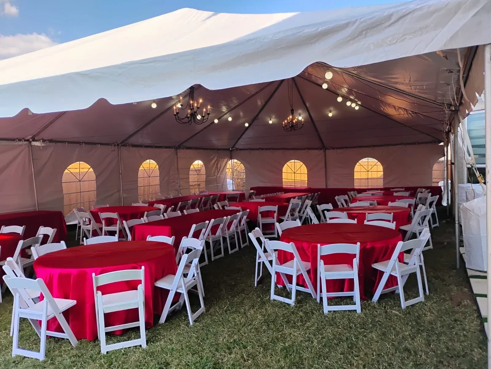 Event tent with red tablecloths, white chairs, and string lights, set on grass.
