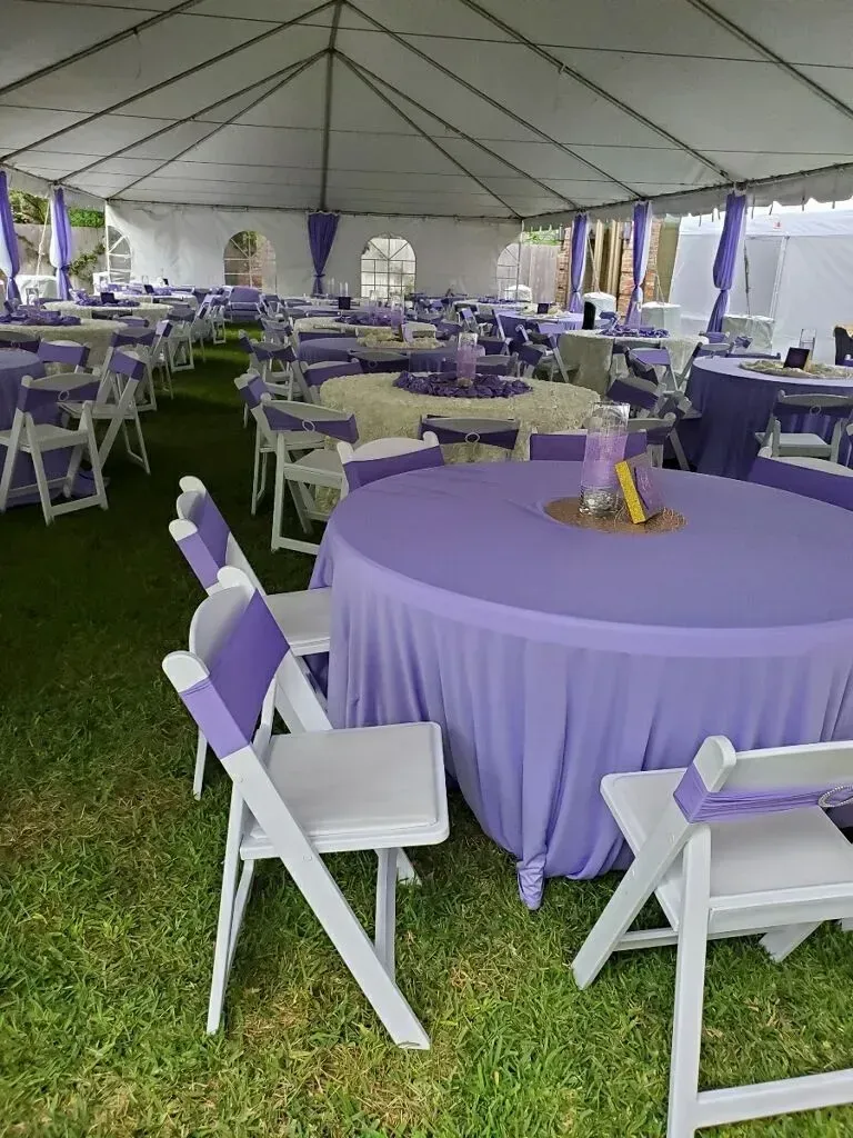 Tables set with lavender linens and white chairs under a white tent for an outdoor event.