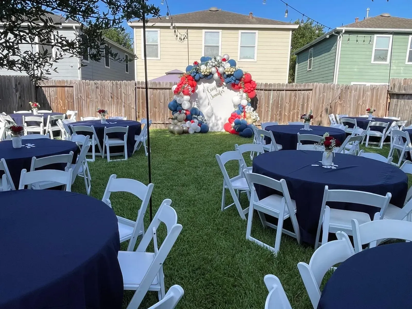 Backyard party setup with round tables, white chairs, navy tablecloths, and a balloon arch in red, white, and blue.