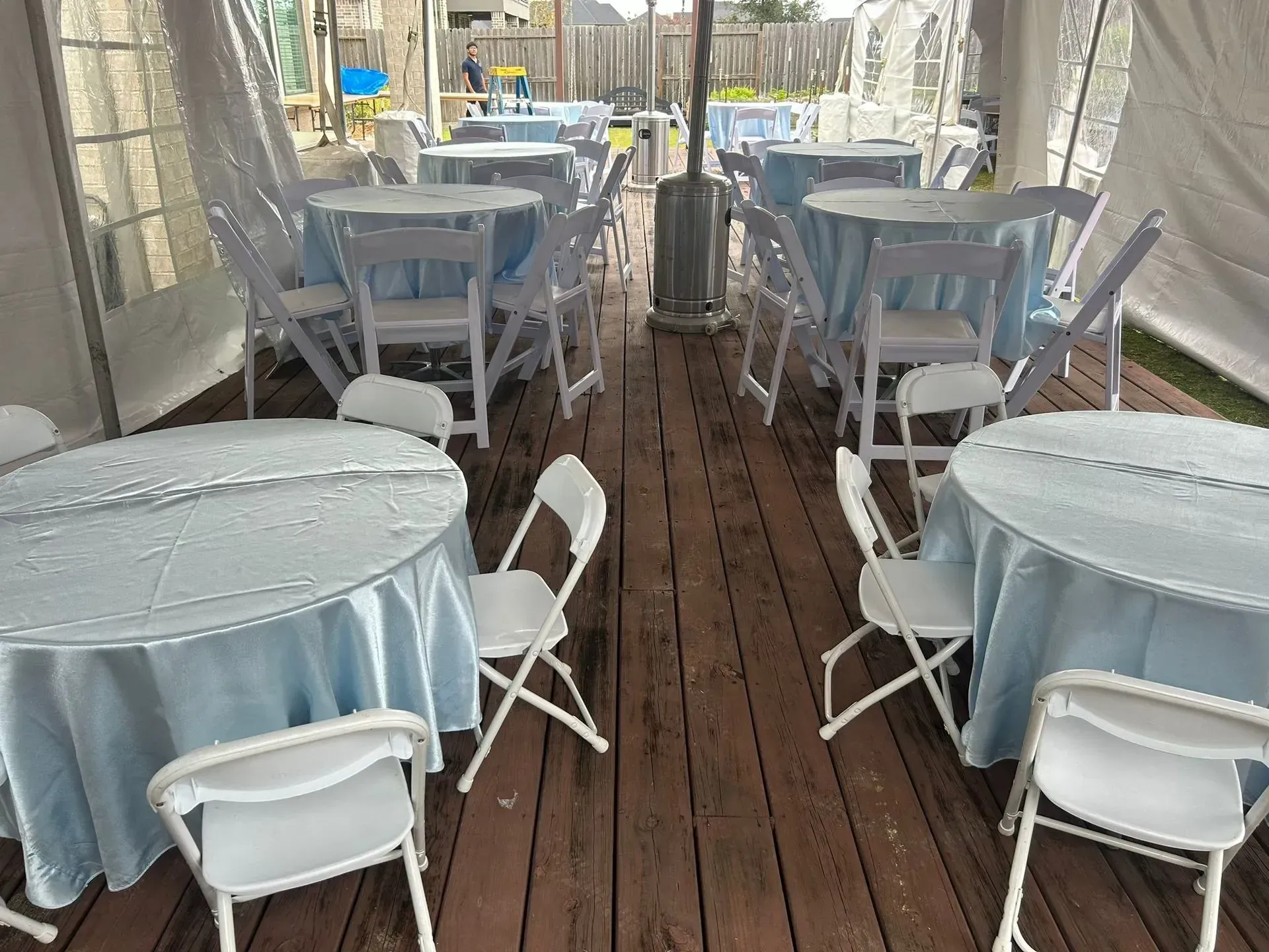 Tented event space with round tables covered in light blue cloths, white chairs, and a patio heater.