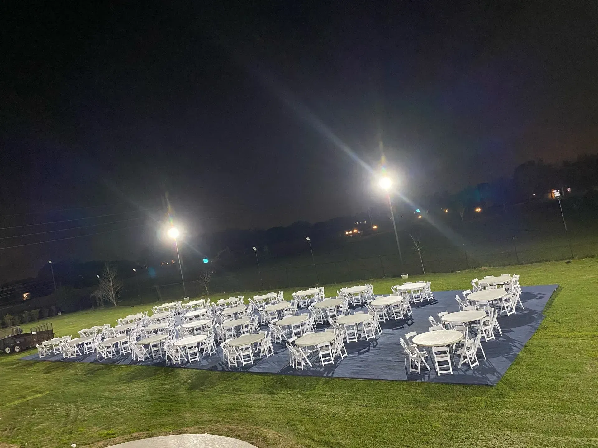 Outdoor dining tables arranged on a blue tarp at night, illuminated by bright lights on a grassy hill.