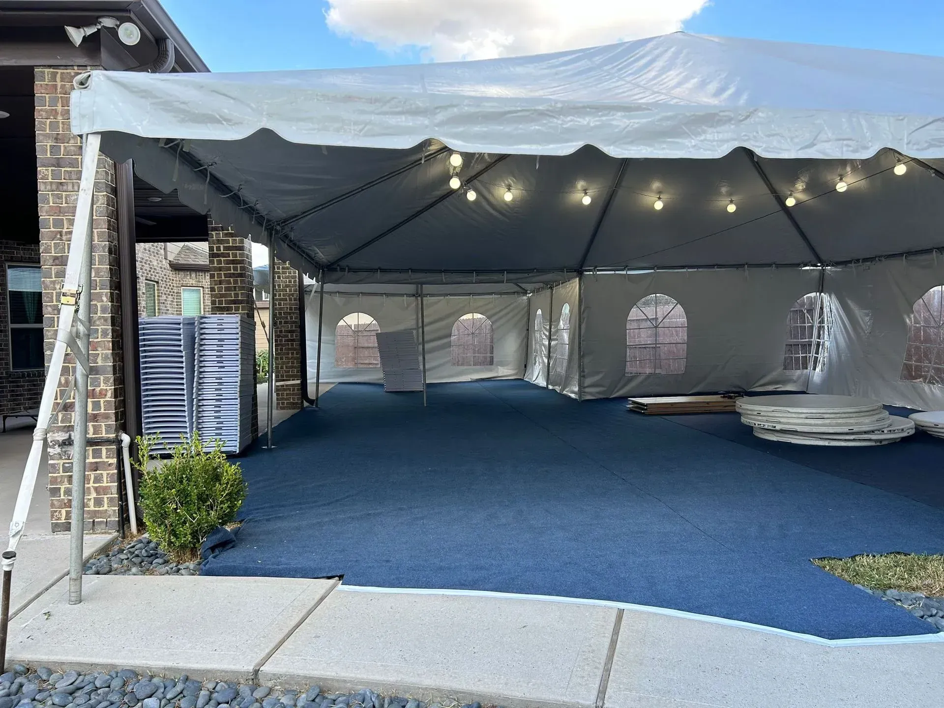 Tent with blue flooring outside a building, with stacks of chairs visible.