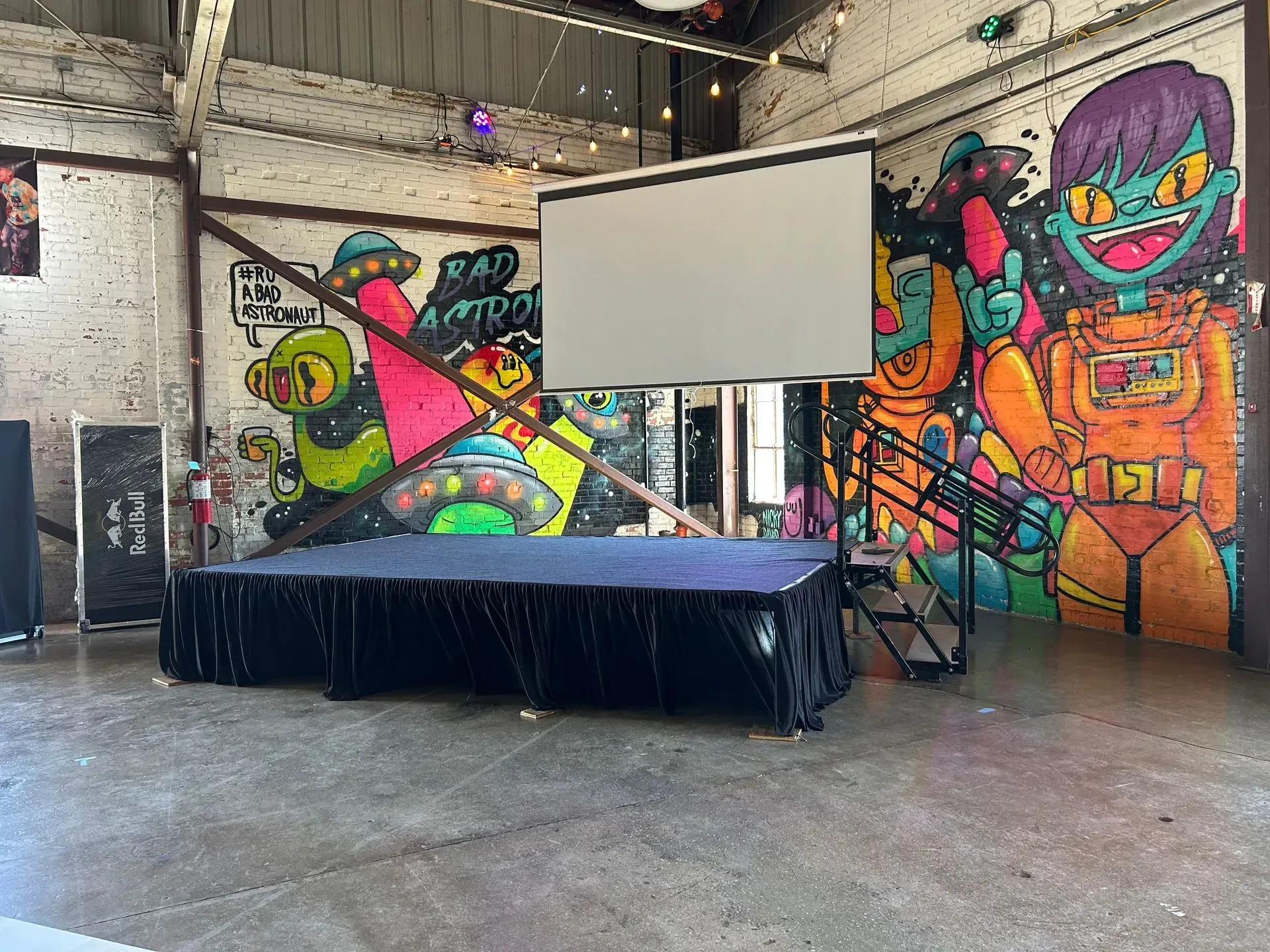 Stage in a venue with graffiti art, projector screen, and a staircase.