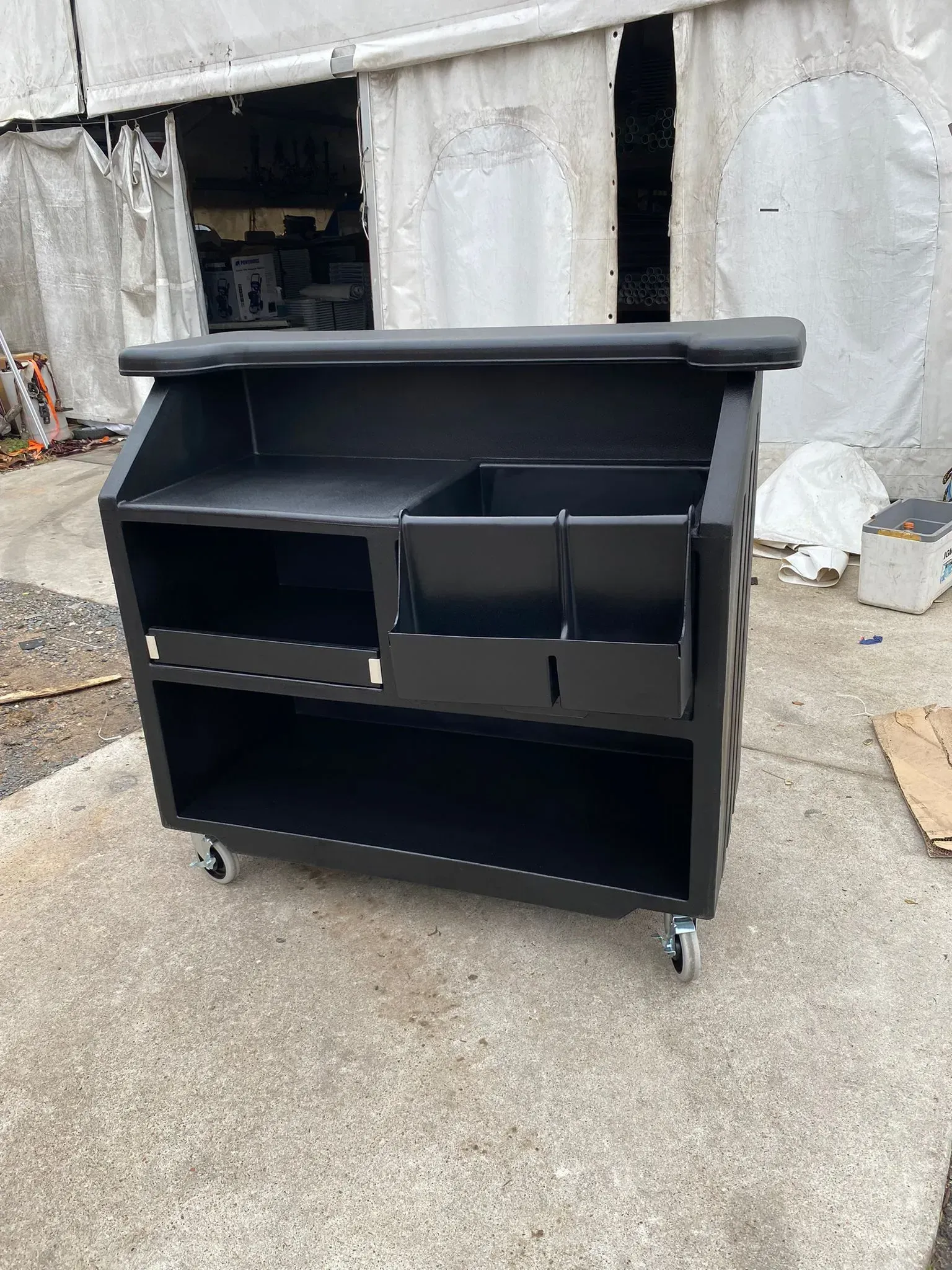Black rolling outdoor bar with shelves, bins, and a top shelf, set outside on concrete.