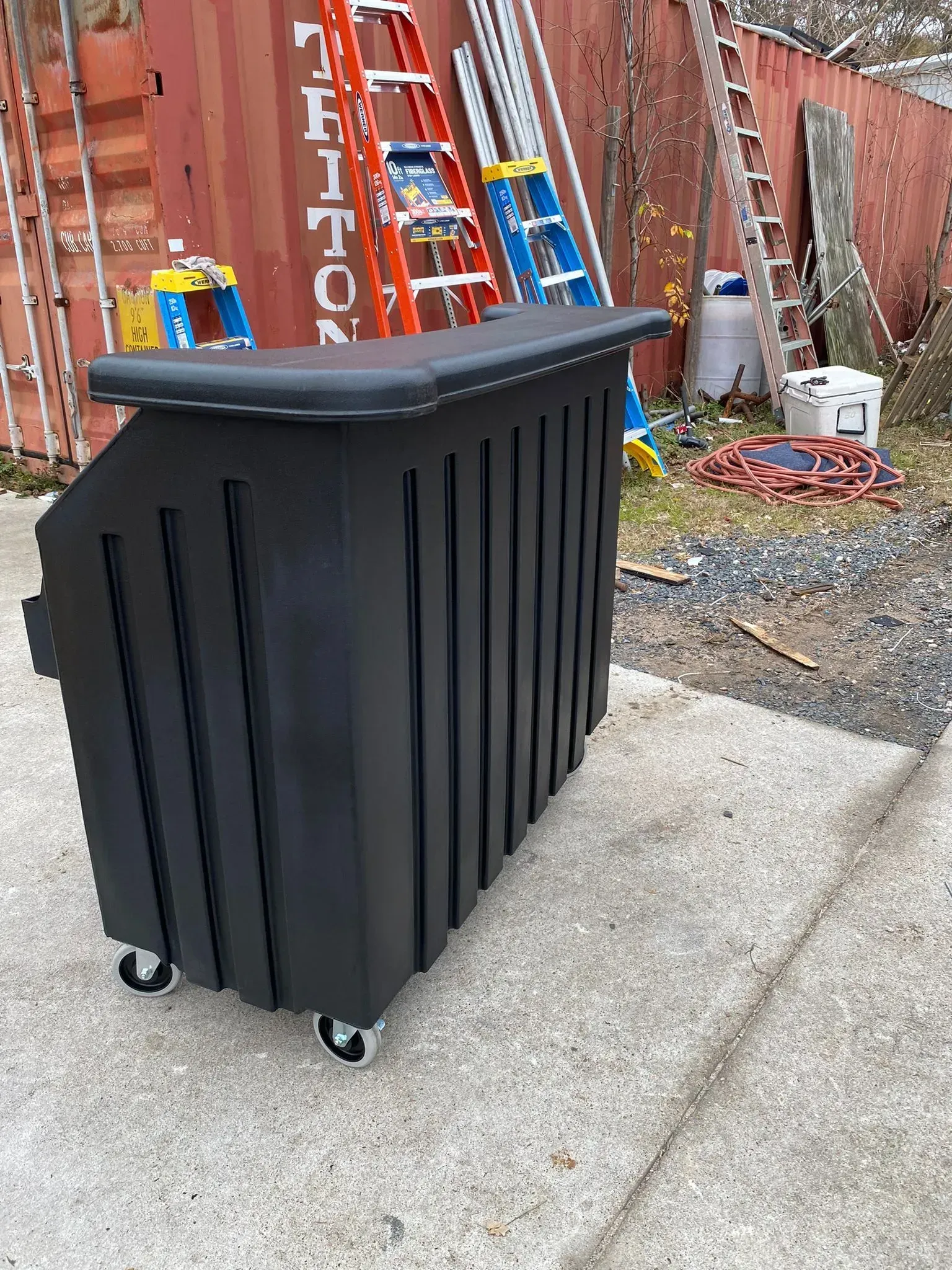 Black portable bar with casters, outdoors. Red shipping container and ladders in background.