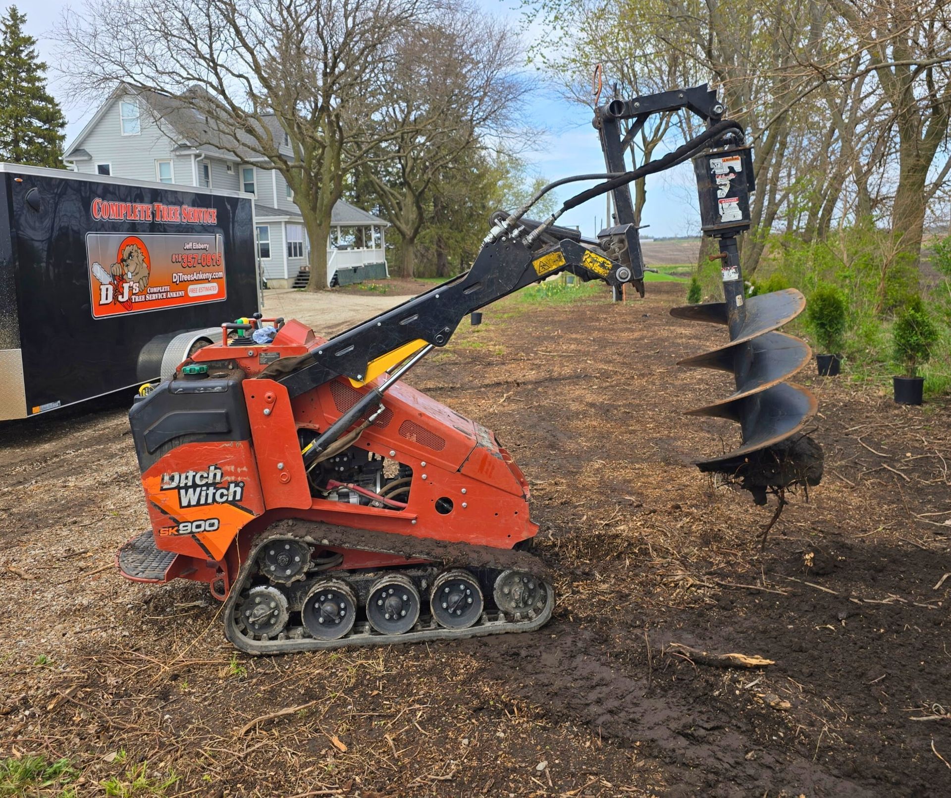 Orange Ditch Witch mini-skid steer with auger drilling holes in dirt. Green trees and black trailer are in the background.