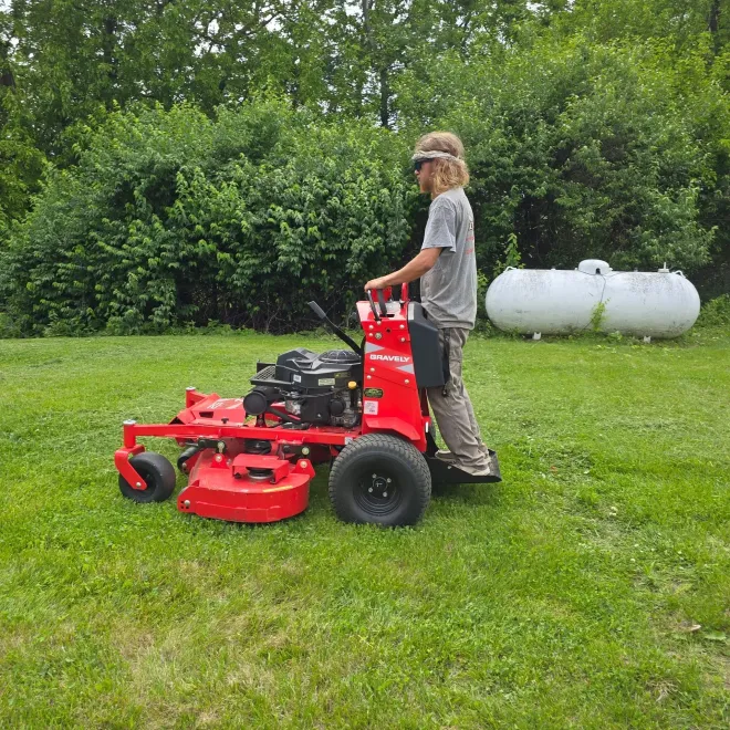 Person operating a red stand-on lawn mower on a grassy yard, with a large white tank in the background.
