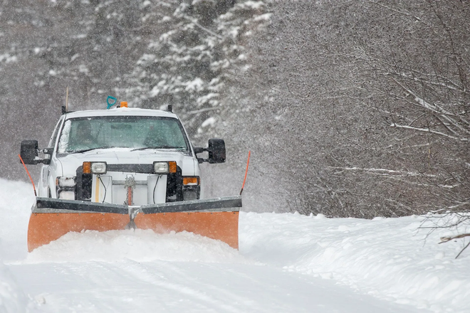Snowplow clearing a snow-covered road during a snowfall; trees line the sides.