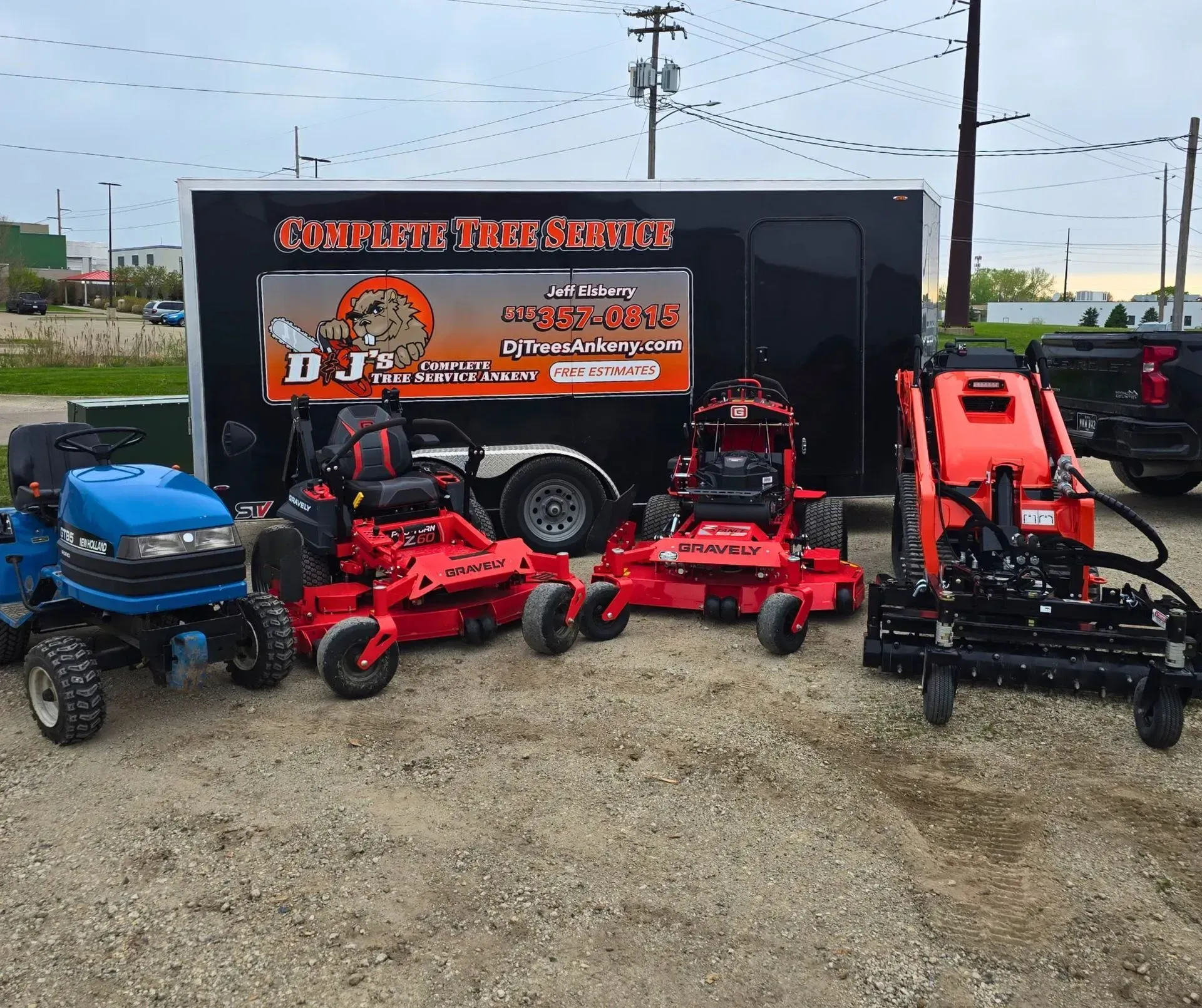 Lawn care equipment: a trailer with business logo, blue tractor, red mowers, and a landscape attachment on a gravel lot.