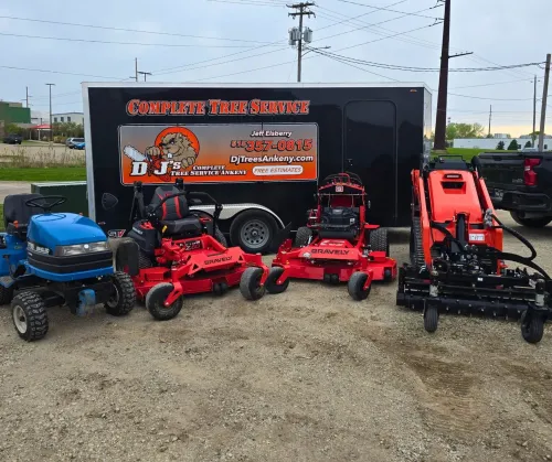 Lawn care equipment: a trailer with business logo, blue tractor, red mowers, and a landscape attachment on a gravel lot.