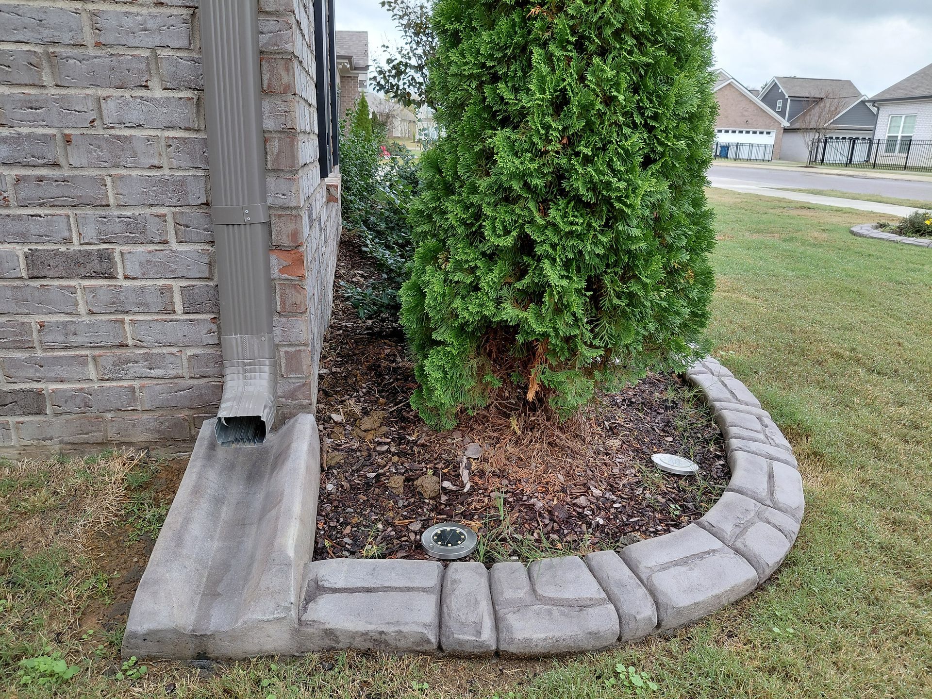 A brick house corner features a downspout extending into a concrete splash block surrounded by decorative stone edging.
