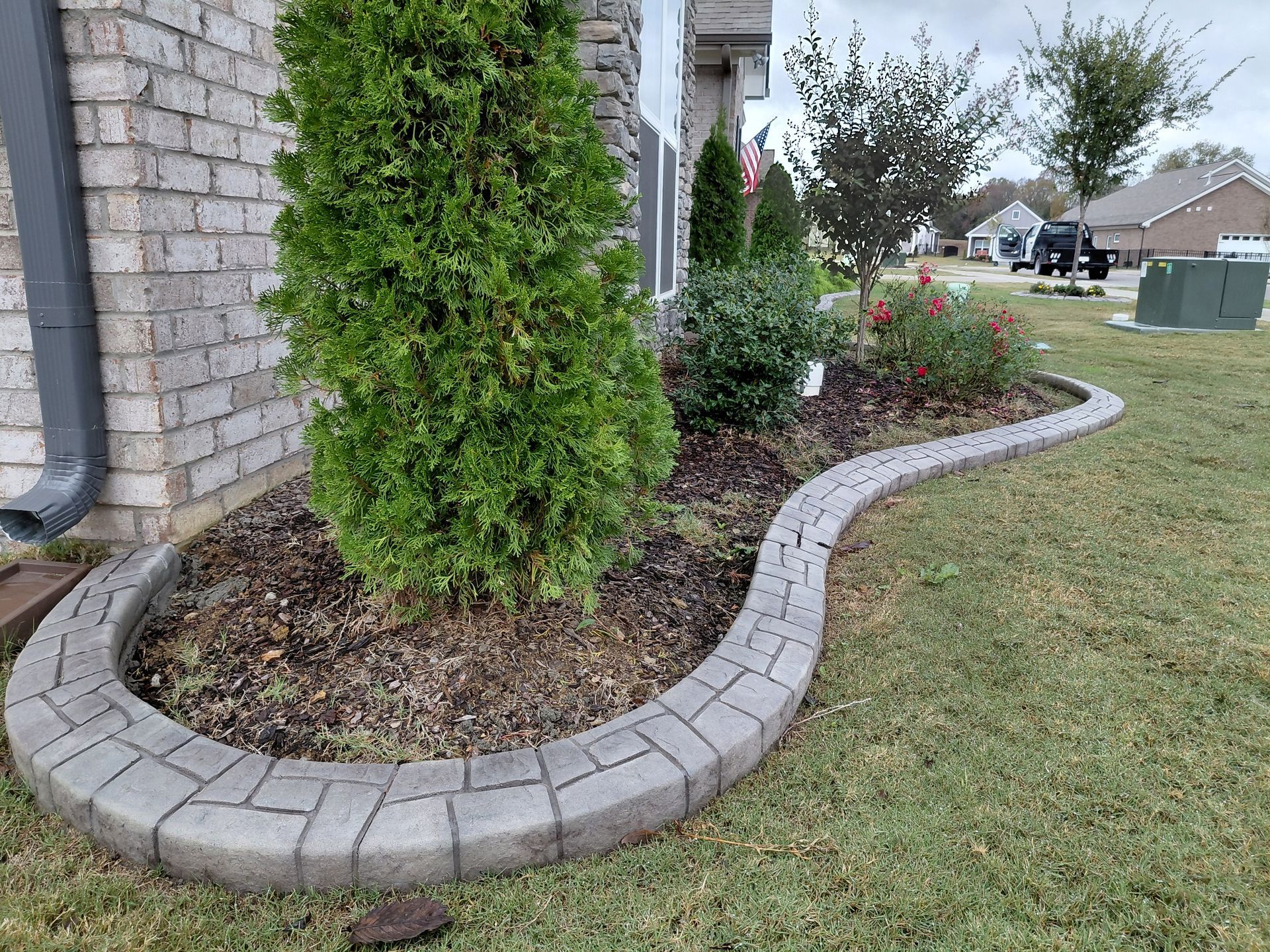 A garden bed with a lush green shrub and small plants, bordered by curved, light gray concrete pavers next to a brick wall.