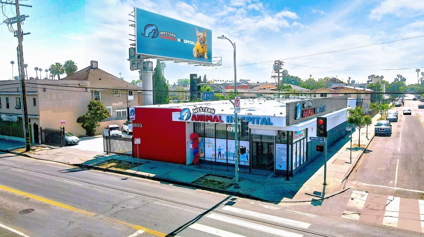 A commercial building with a large red wall, sign and billboard on a sunny street with cars.