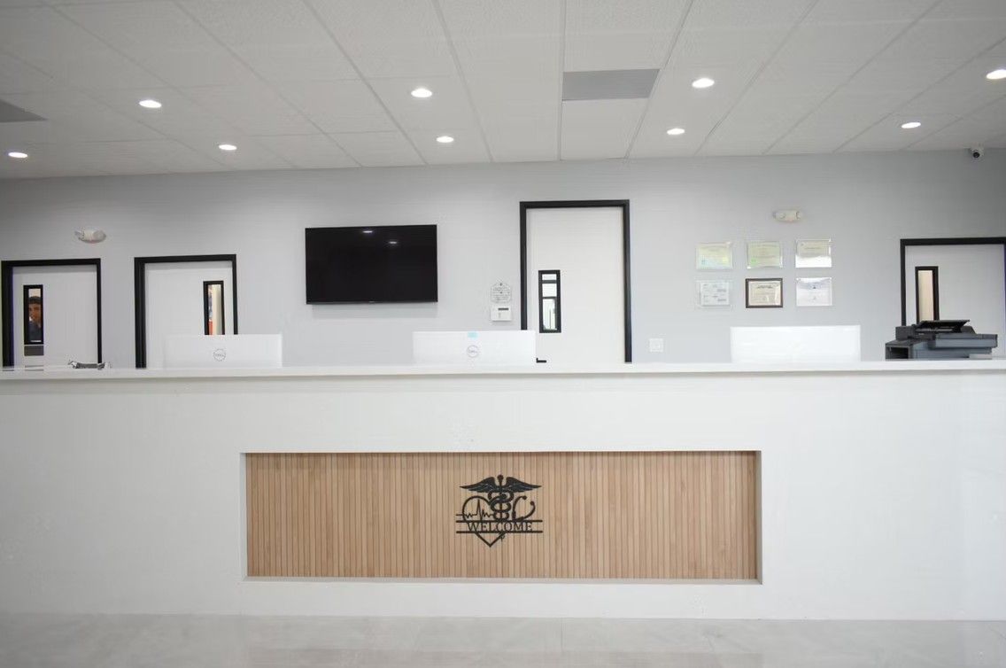 Reception desk in a medical office; white counter, wood panel, TV, doors, diplomas, and logos against a gray wall.