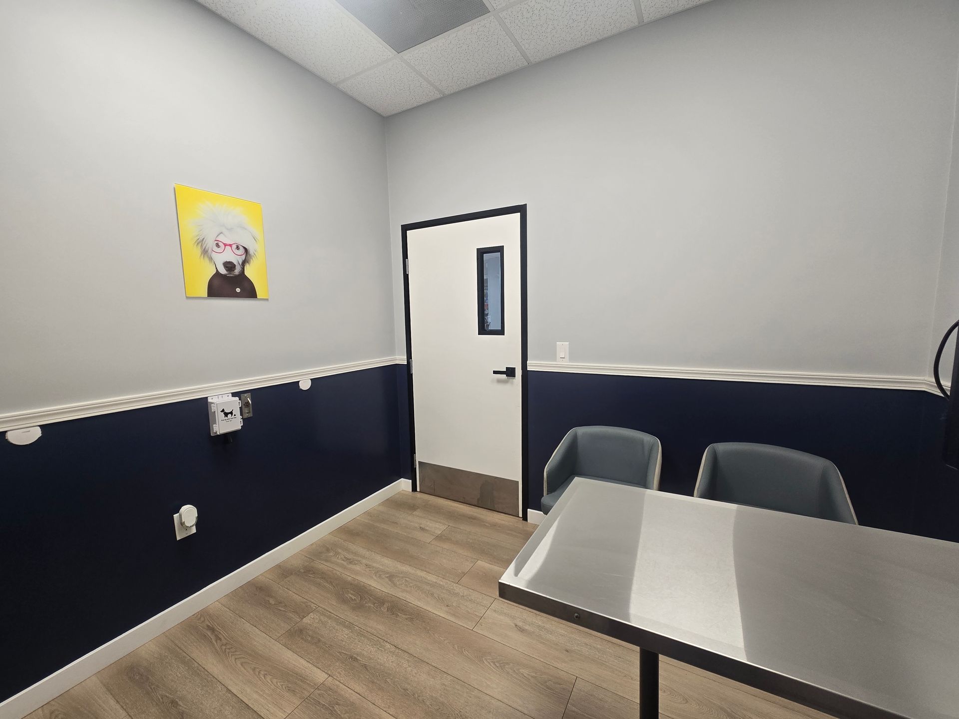 A small room with light wood floors, a stainless steel table, two chairs, and a portrait. A door is on the back wall.