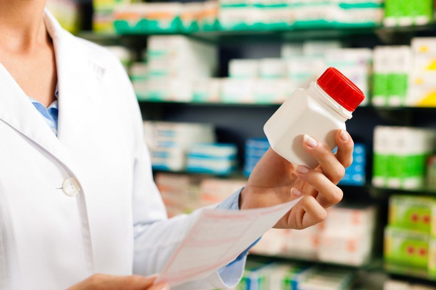 Pharmacist in white coat, holding prescription and pill bottle with red cap in a pharmacy.