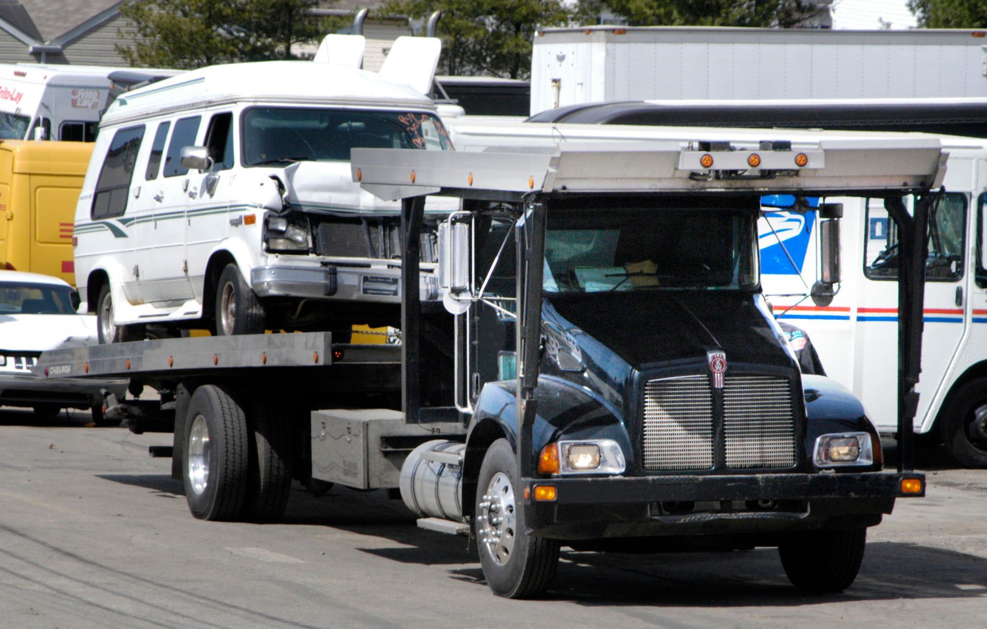 Black tow truck carrying a white van on a flatbed. Outdoor, daytime.