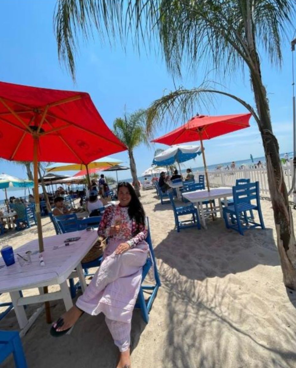 Woman relaxing at a beach table under red umbrellas, with palm trees and ocean visible in the background