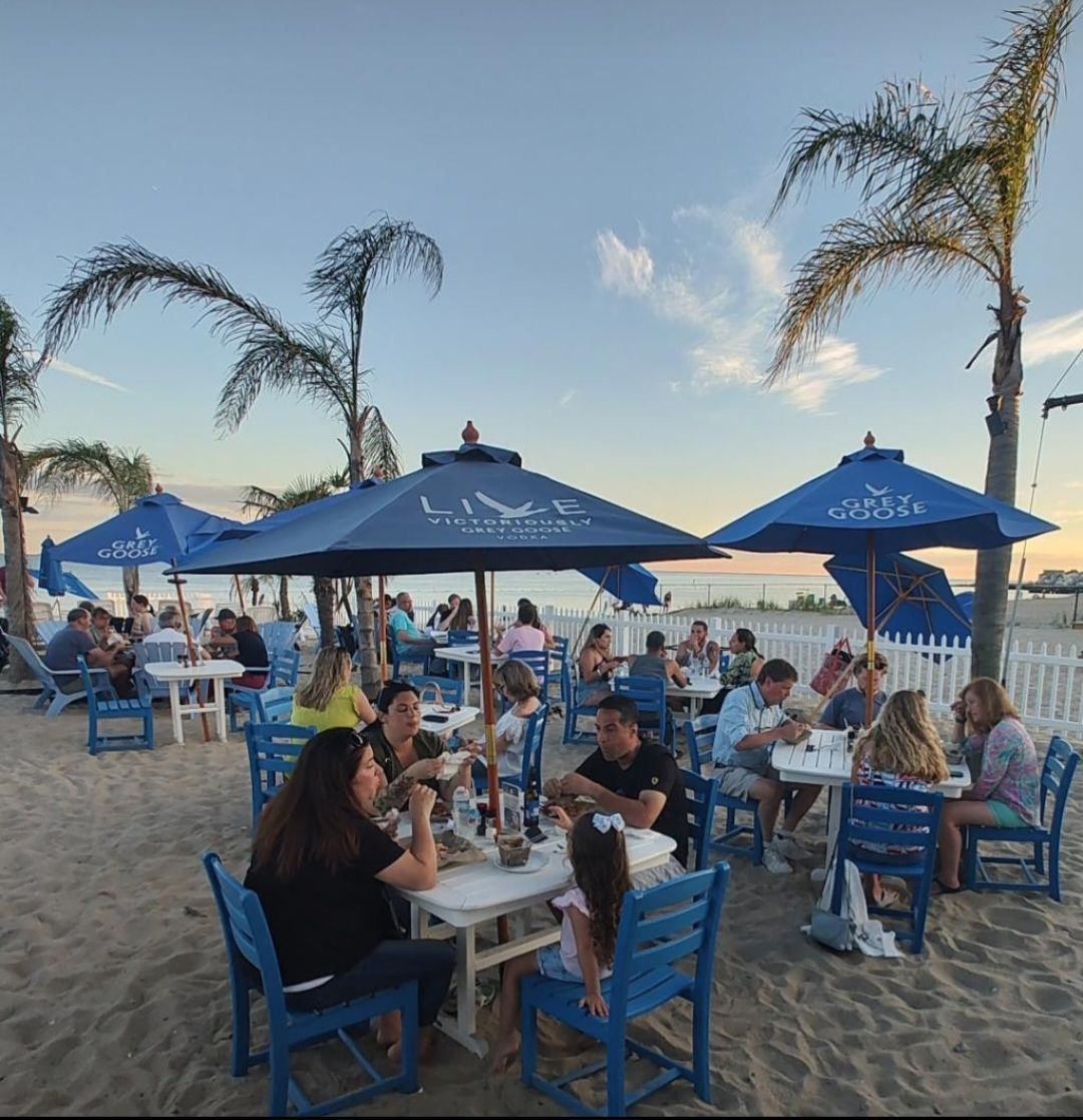 People dining at blue umbrellas on a sandy beach at sunset, with palm trees and the ocean in view.