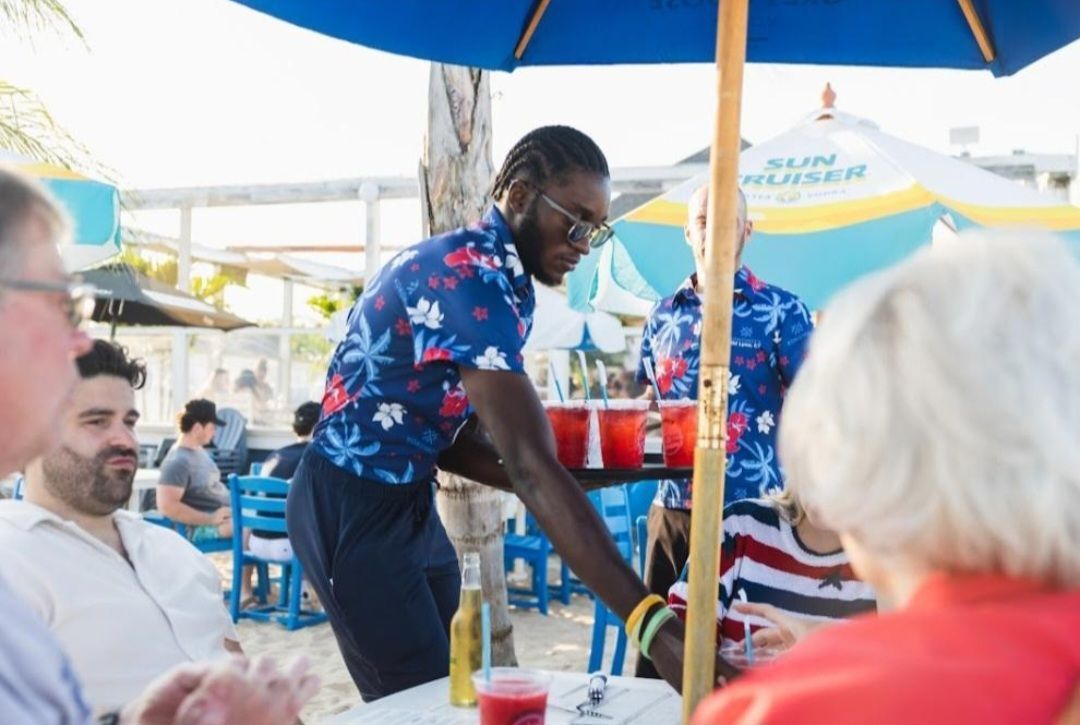 Waiter serving drinks at an outdoor café under a blue umbrella, with seated patrons nearby