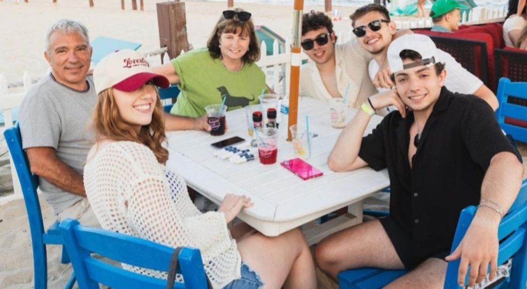 Group of friends smiling at an outdoor table, wearing casual clothes and sunglasses, with drinks and a blue chair.