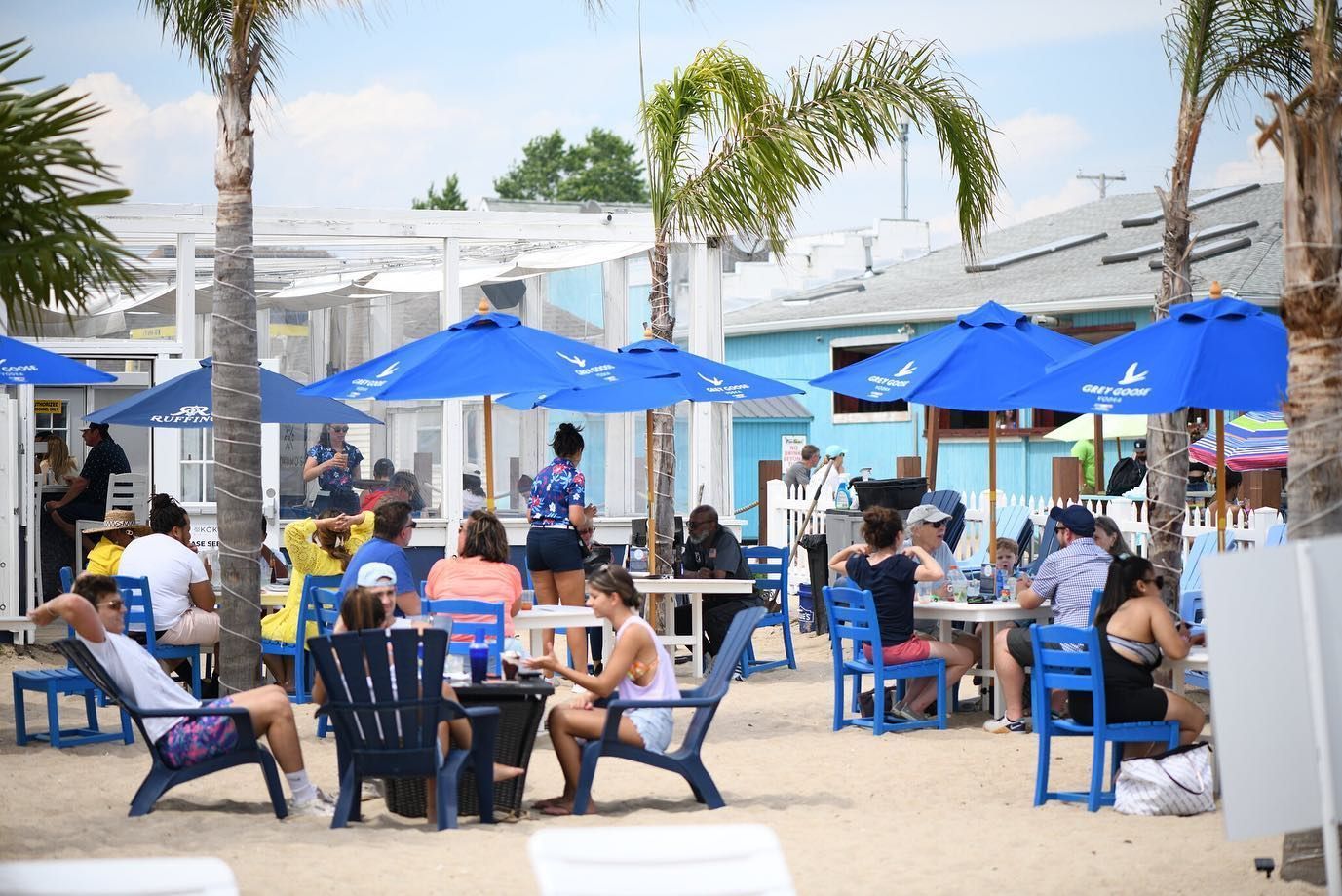 Beachside café with blue umbrellas, people seated at tables, and white lounge chairs near the water