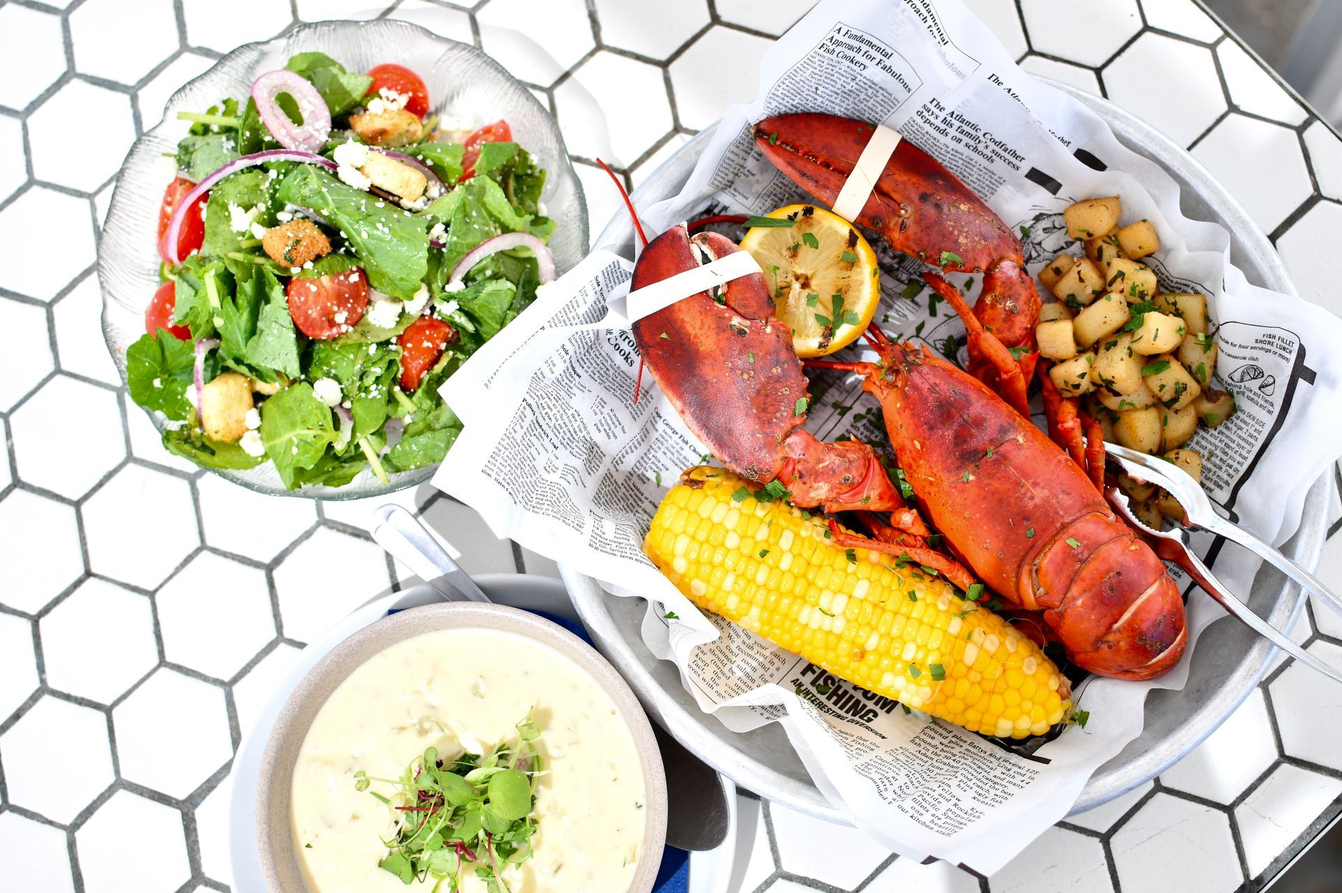 Seafood boil with lobster, corn, potatoes, salad, and soup on a white tiled table