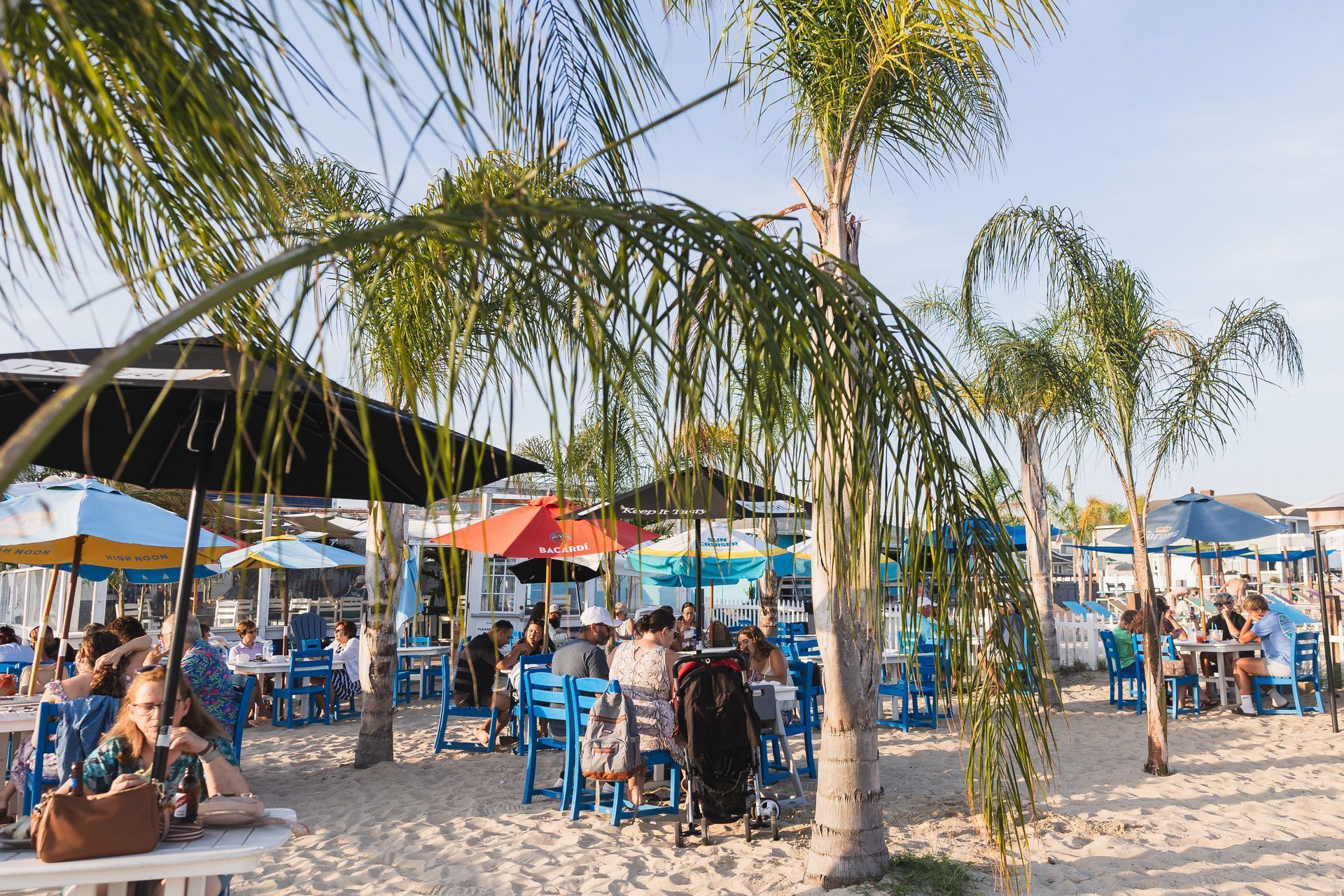 Beachside restaurant with blue tables and chairs, palm trees, and colorful umbrellas. People sit and dine on the sand.