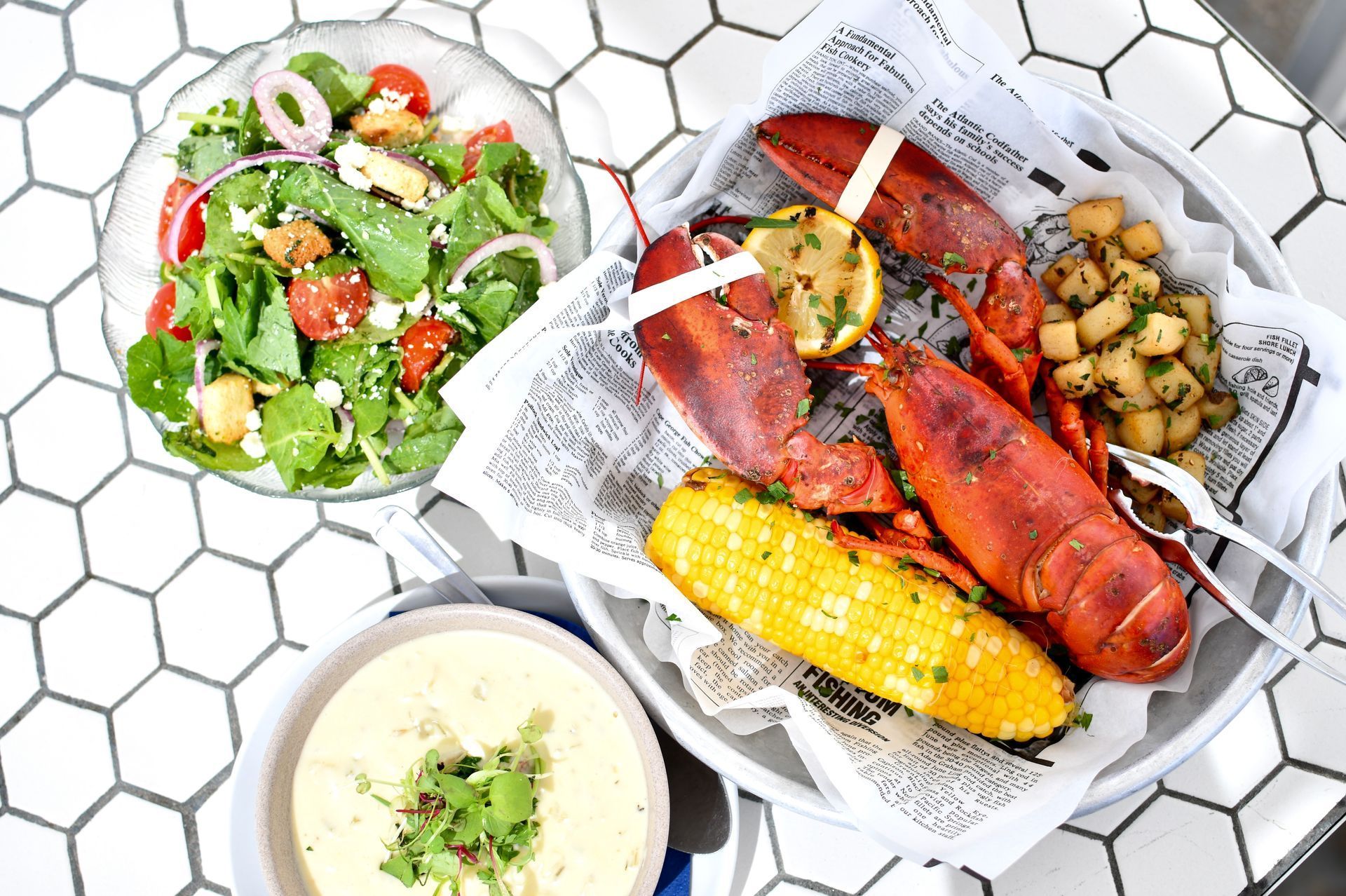 Lobster dinner with salad, corn on the cob, potatoes, and soup on a white table.