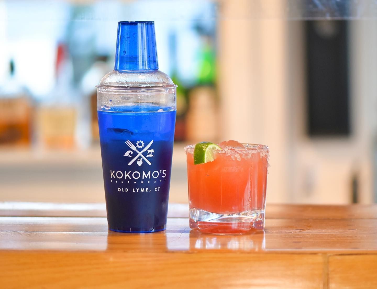 Blue cocktail shaker next to a red cocktail with lime garnish, on a bar.