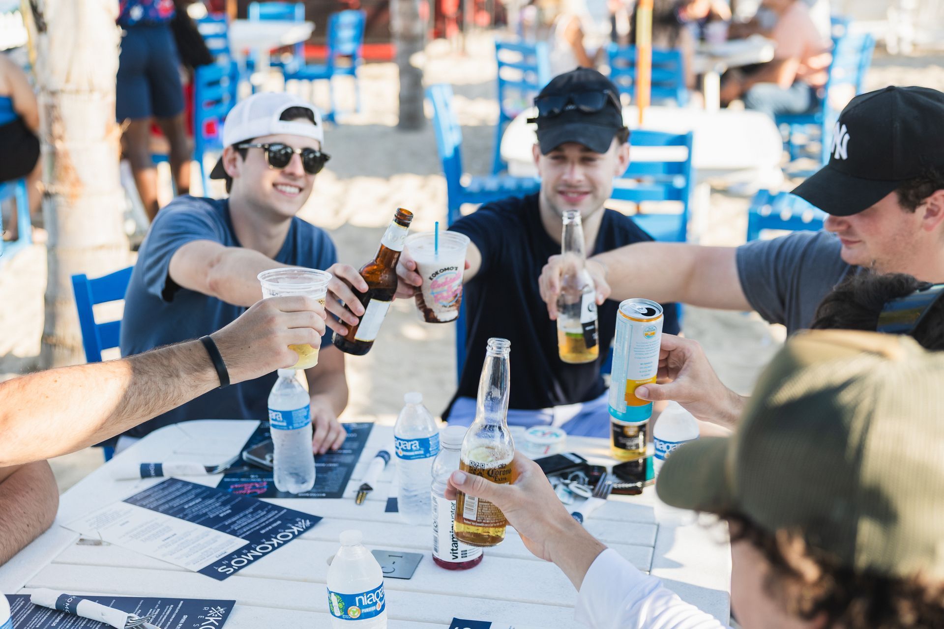 Friends toasting beer bottles and plastic cups at an outdoor table.