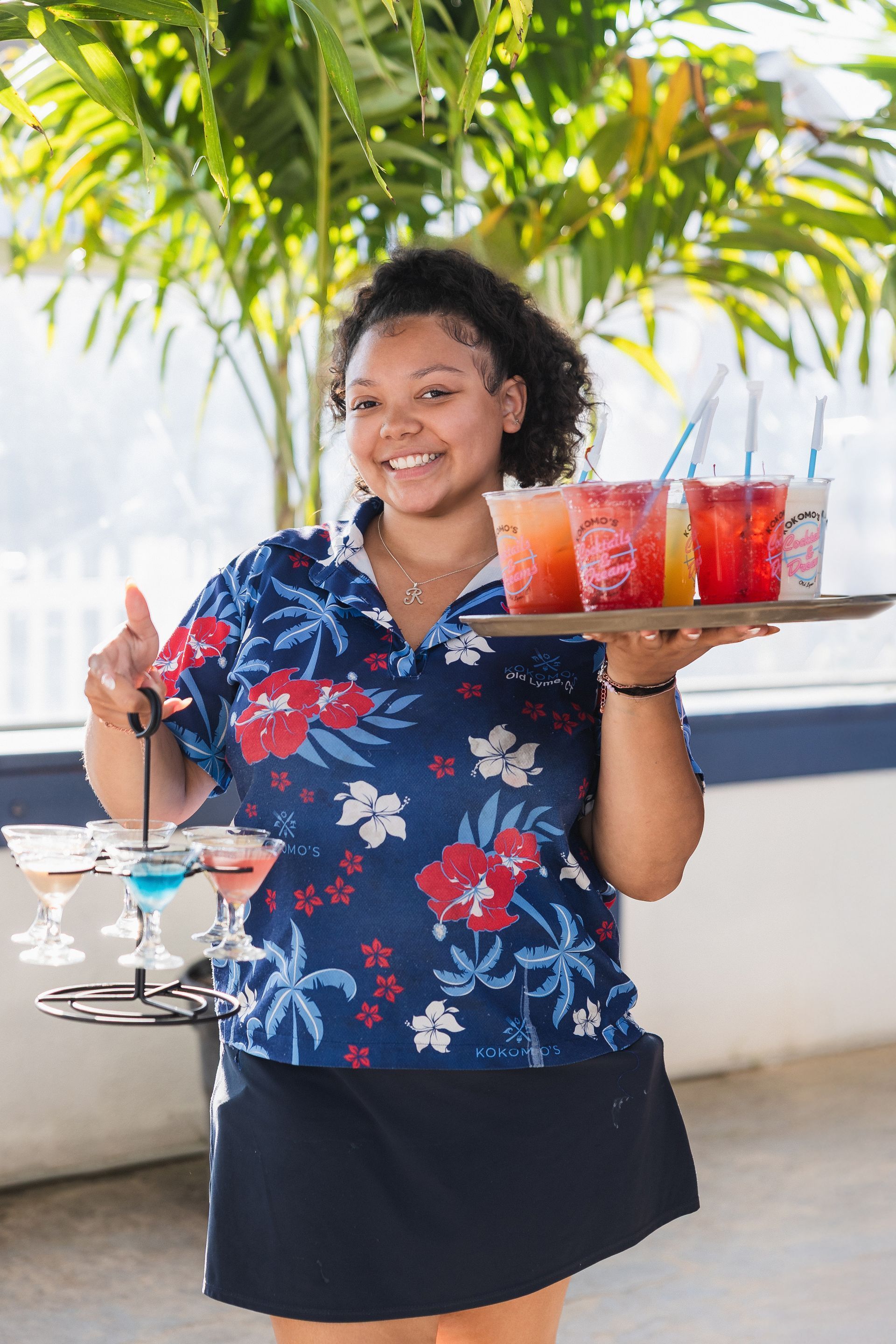 Smiling woman in a floral shirt carries trays of drinks outdoors.
