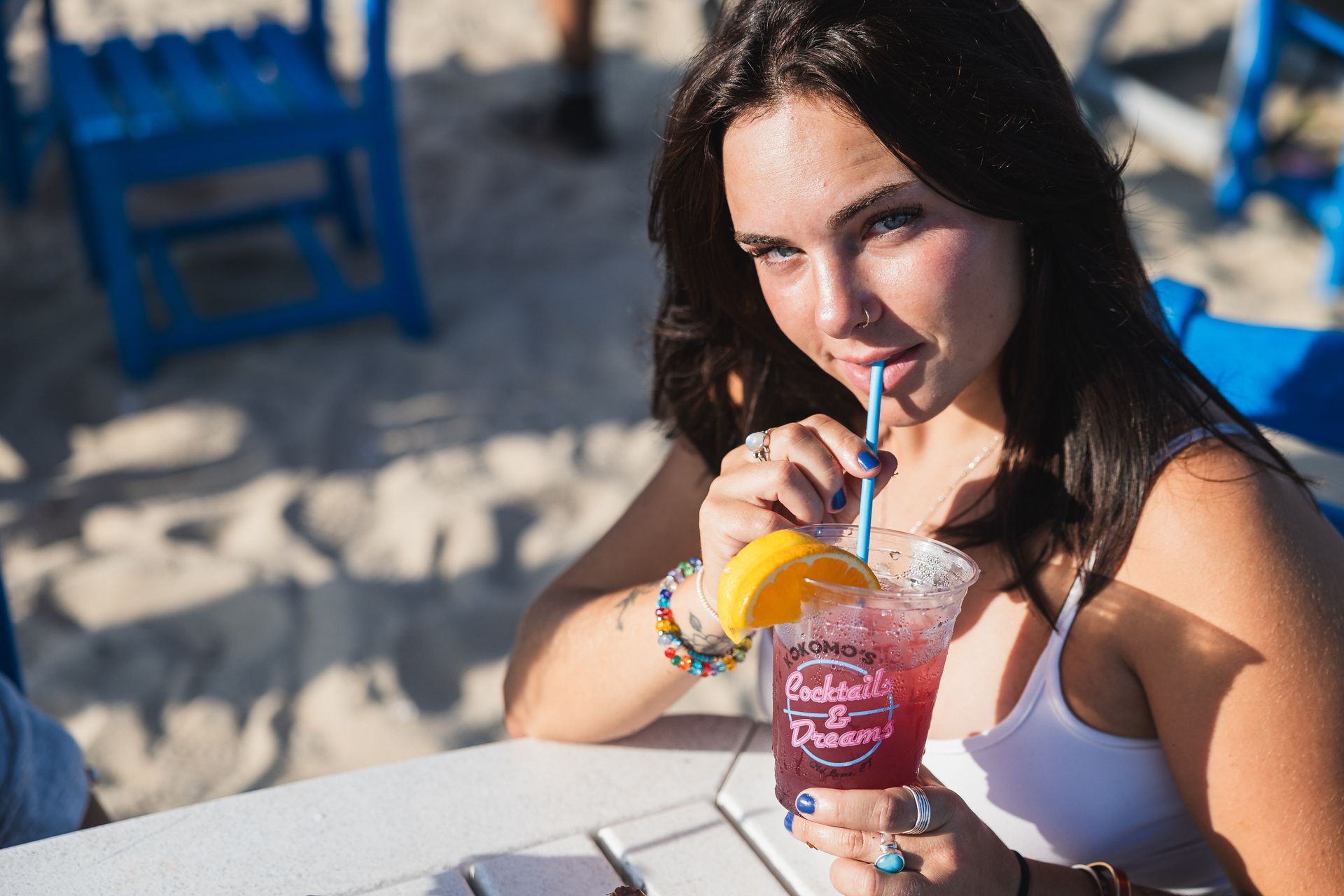 Woman sipping a red cocktail with a straw at an outdoor table on a beach.