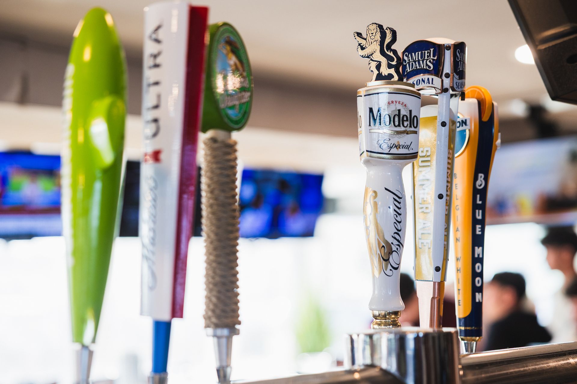 Beer taps lined up at a bar, various brands, bright colors, blurred background with people.