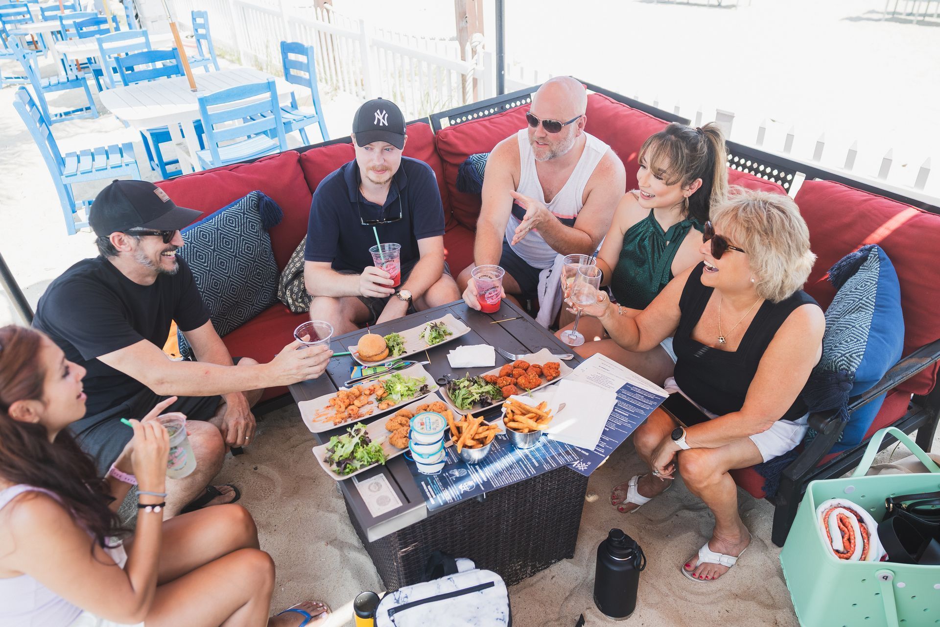 Group of six people eating food and drinking on a red outdoor couch, with a blue waterfront in the background.