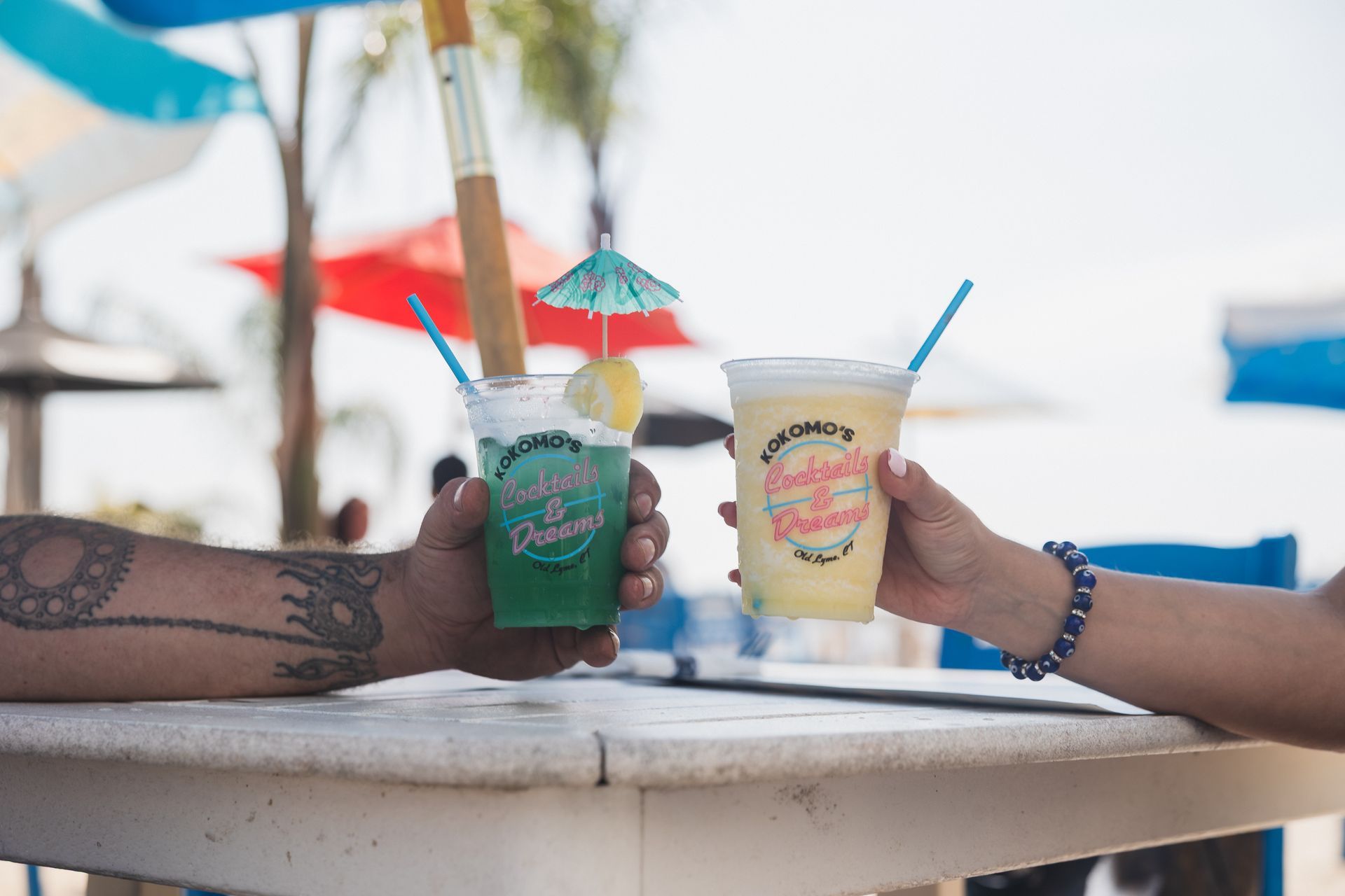 Two people toasting colorful tropical drinks at an outdoor table.