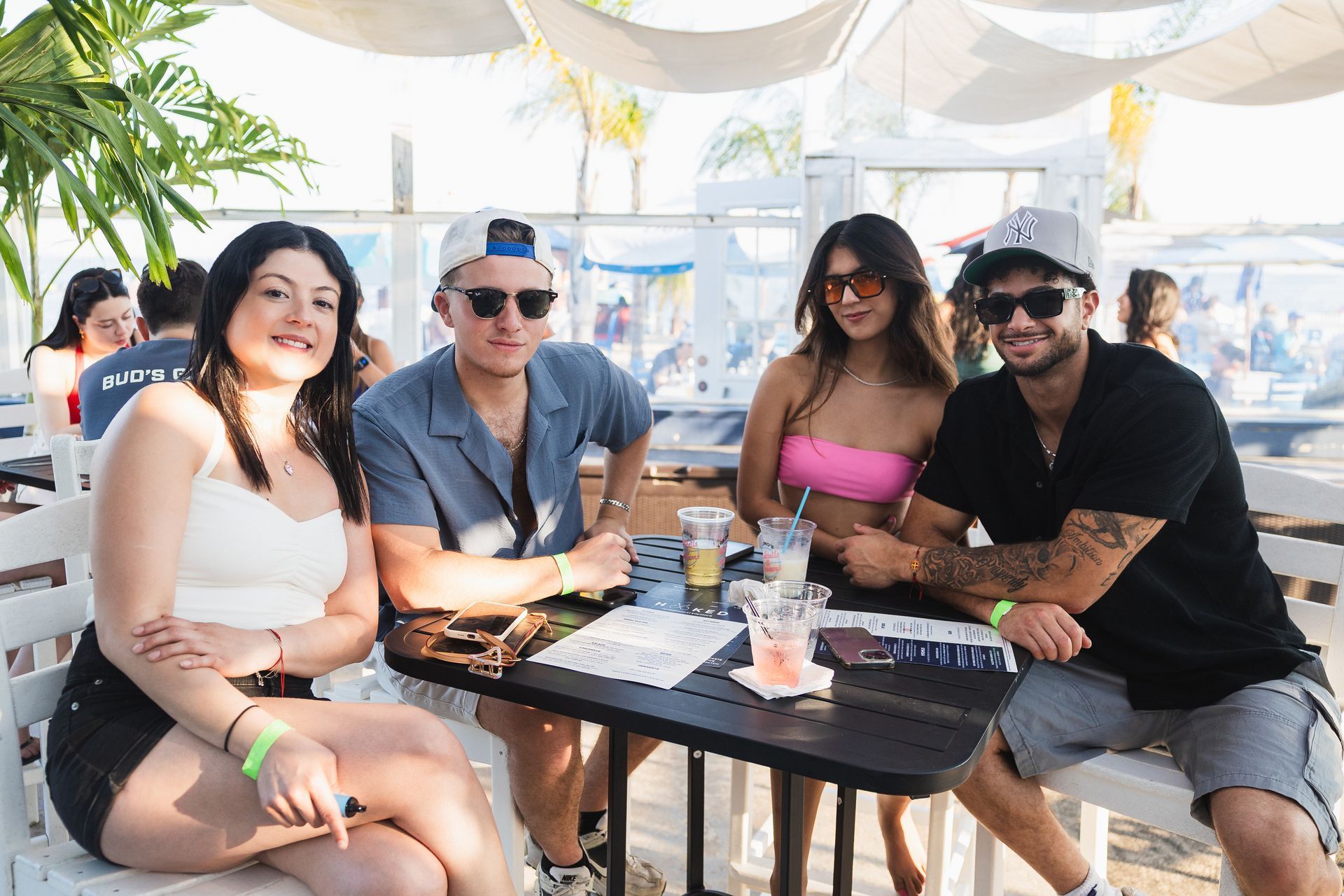 Four people seated at an outdoor table, smiling, with drinks. Beach setting, bright sunlight.