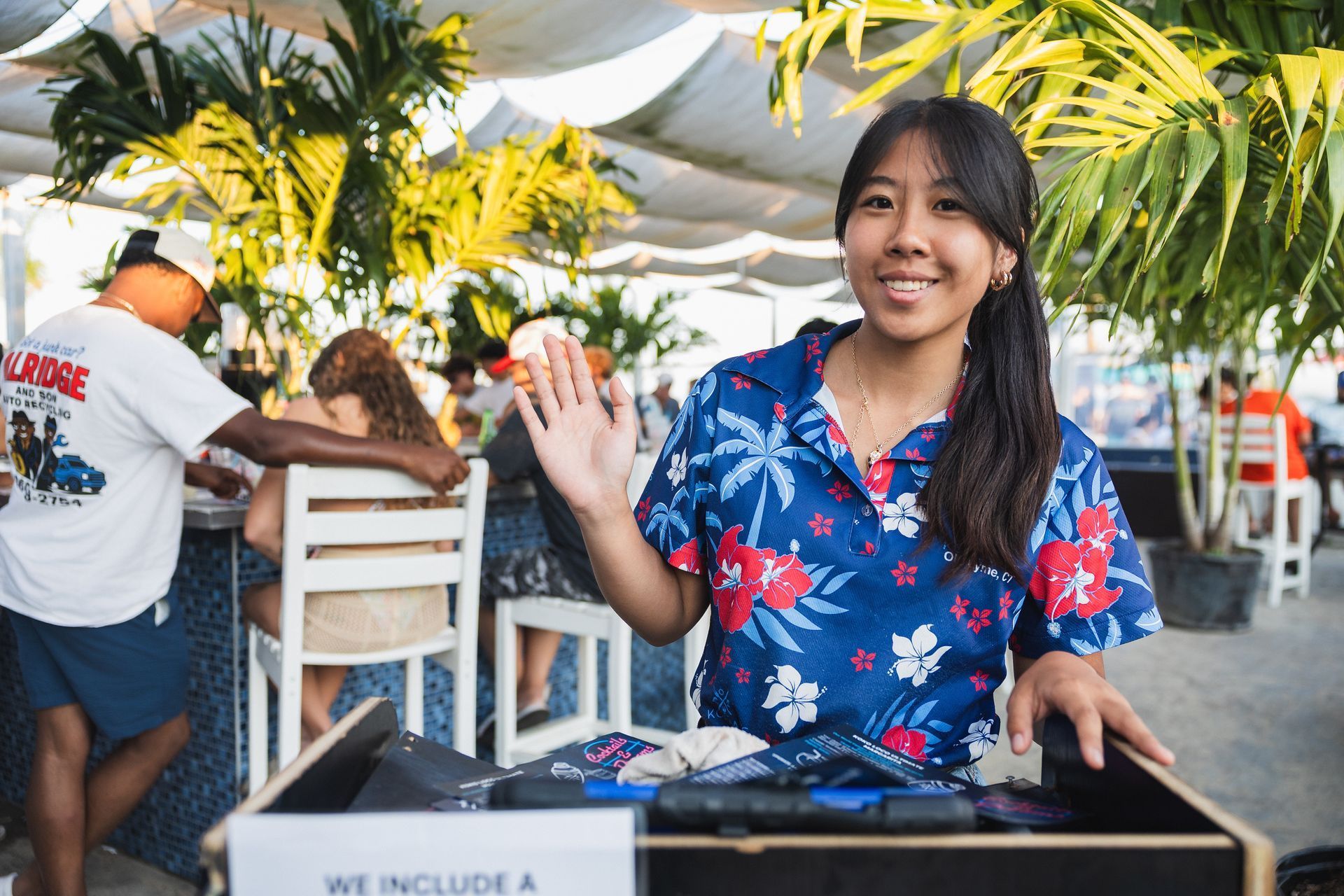 Woman in floral shirt waves, standing near a box at an outdoor bar.