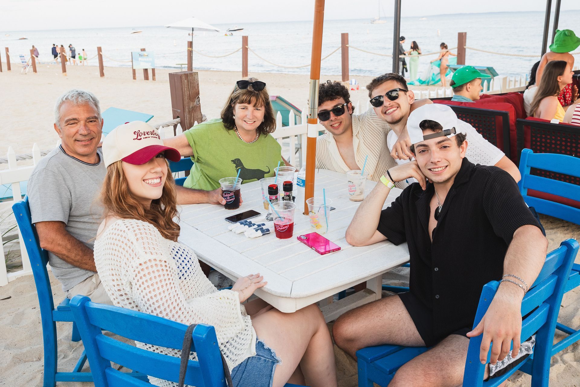 Group of people smiling around a table on a beach, under a parasol; drinks, sunglasses, summer.